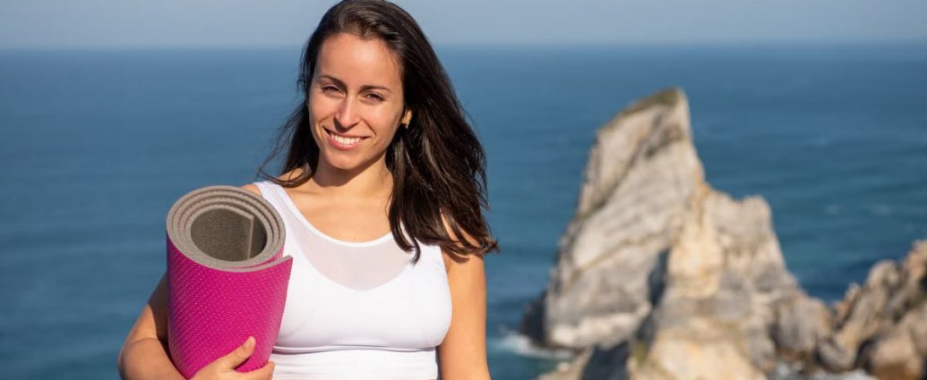 Smiling woman with yoga mat in sportswear by oceanic cliffs in Portugal.