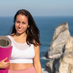 Smiling woman with yoga mat in sportswear by oceanic cliffs in Portugal.