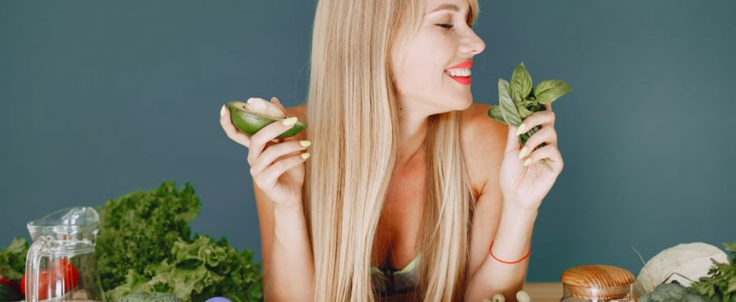 A smiling woman holding avocado and basil, surrounded by fresh vegetables and fruits for healthy eating.